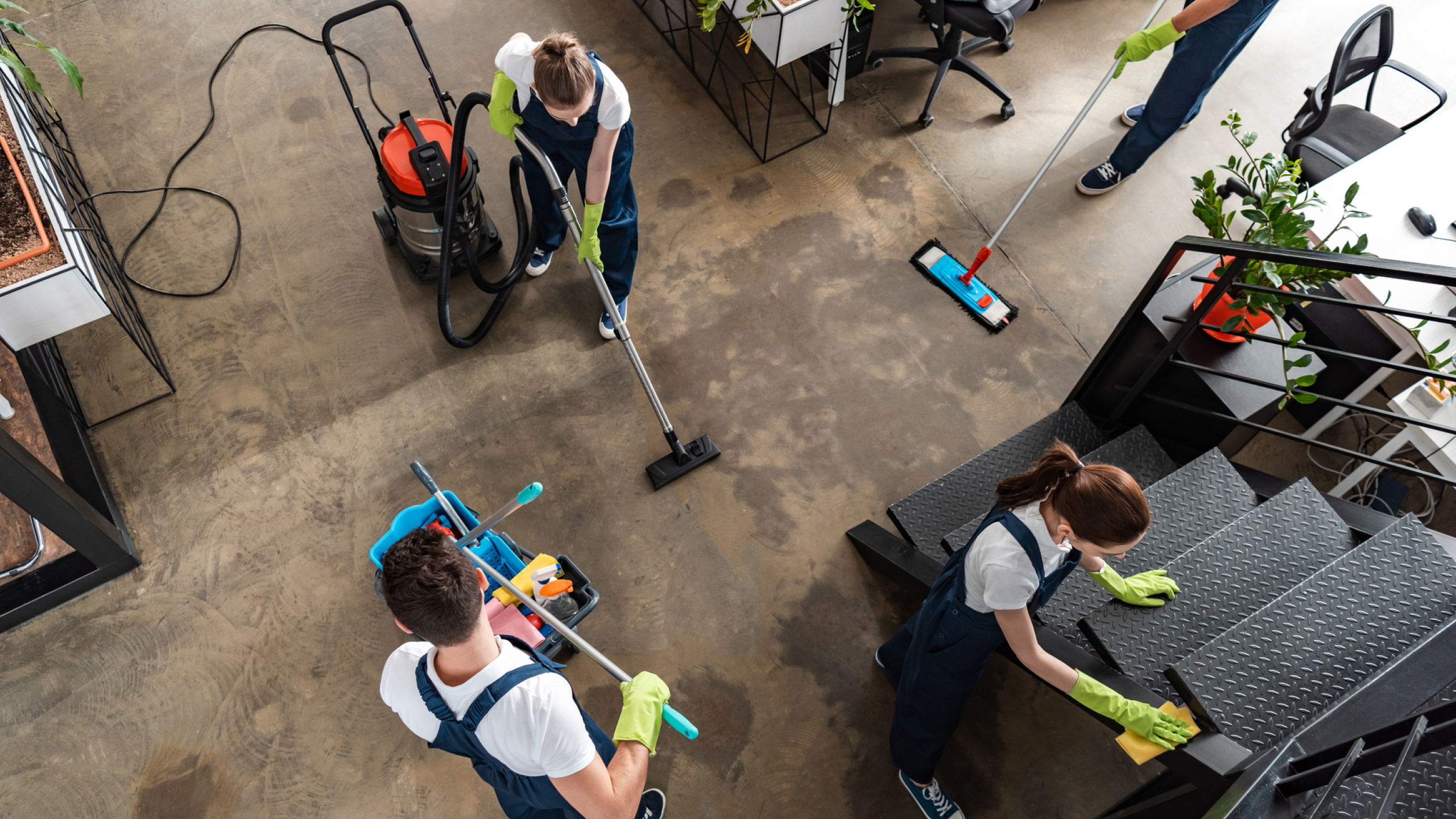 Two individuals are cleaning a spacious area with a vacuum and mop, while another uses a pressure washer nearby. They are all wearing matching uniforms and gloves, with various cleaning equipment and plants visible around the office setting.